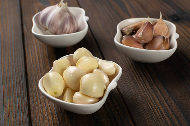 Small white bowls with peeled and unpeeled garlic on a wooden table.